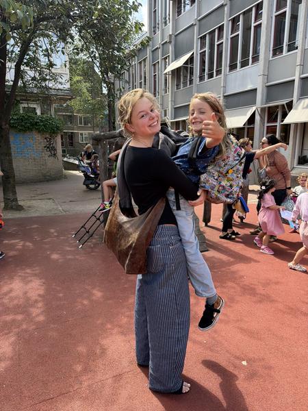 A woman holds a young child, both smiling brightly at the camera, in a sunny outdoor schoolyard at Lastageweg, Centrum, Amsterdam